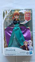 Front view of sealed Anna doll in original packaging, showing the doll wearing a teal and purple dress, with red hair in braids. Box indicates musical feature with song 'Into the Unknown.'