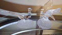 Two clear crystal bird figurines on a glass table, one with wings spread and the other a long-tailed pheasant with brand sticker