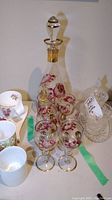 Overall view of the glass decanter, six matching glasses, and additional items on table, focusing on decanter and glasses painted with floral designs.