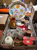 Top view of a box containing various ceramic and wooden items including candlestick holders, honey container, small pitcher, mugs, and a colorful round tray.