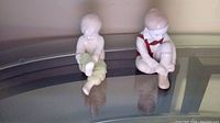 Two white ceramic child figurines on glass surface, one tying shoe wearing red bowtie detail, one sitting with legs crossed.