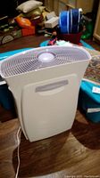 White plastic filtration humidifier with vented horizontal top and recessed handle on front. Unit standing on wood floor with various household items in background.