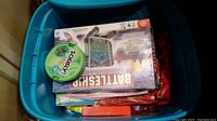 Blue plastic bin containing multiple board games including Battle Ship and Sorry!, puzzles, and toy figures.