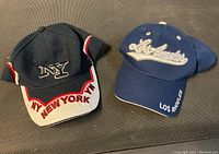 Two baseball caps: one navy blue cap with NY logo and white-red visor accents, one navy blue cap with Los Angeles text embroidered on front and side.