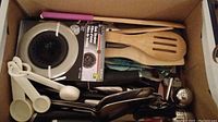 Photo shows a box containing multiple kitchen utensils including wooden spoons, metal utensils, KitchenAid cutters in packaging, white plastic measuring spoons, metal sink strainer and bottle opener.