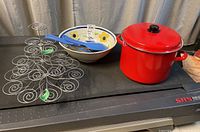 Photo showing red cooking pot with lid, floral bowl, blue kitchen utensils inside the bowl, and metal cupcake stand arranged on black surface
