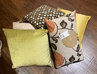 Five pillows stacked on wooden floor showing different styles and sizes, with floral and geometric patterns as well as solid colors.