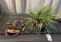 Photo showing one faux plant with long grass-like green leaves in a black pot, wristwatch with metal band, yoga mat and decorative fabric pouch with gold and red intricate patterns.