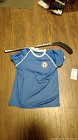 Chelsea Football Club blue shirt laid flat on wooden floor next to Bauer hockey stick.