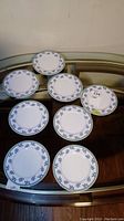 Eight round porcelain side plates arranged on a glass-topped table, showing their blue leaf patterned rims.