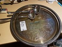 Photo showing circular silver plated tray with embossed detailed design and two silver ladles on top.