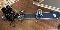 Snowboard laying flat on the floor with blue and white bindings and black snowboard boots resting beside it.
