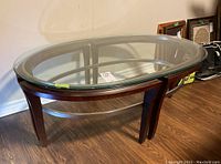 Front angled view of an oval coffee table with clear glass top and wooden legs, showing the metal frame underneath the glass.