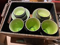 Top-down view of green and black ceramic tea cups arranged inside a woven box, showing both small and large sizes with matching saucers.