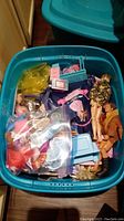 A teal plastic storage bin filled with various Barbie dolls, Barbie accessories, and toys, some items packed in clear plastic bags.