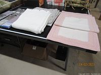 Two bath mats displayed on a table; one white with a pink border, the other white/cream colored, stacked on left.