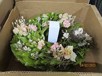 Zoomed out view showing one spring wreath with large green leaves and pastel artificial flowers inside a cardboard box.