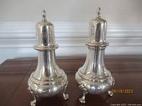 Two silver pepper shakers side by side on wooden surface showing overall shape, feet, and monograms.