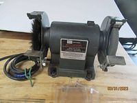 Front view of Craftsman bench grinder showing the body with model and warning labels, tool rests, grinding wheels and wire brush wheel mounted. The power cord is visible coiled next to it.