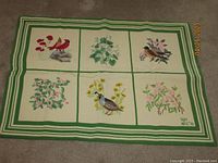 Full view of entire needlepoint rug laid on carpet, showing six different panels with bird and flower designs and striped green and white border.