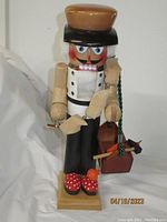 Front view of a 15-inch tall wooden nutcracker, showcasing a shoe maker with a brown hat, black and white attire, red shoes, white beard, holding a wooden shoe and toolbox with shoe-making tools.
