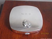 Closed rectangular sterling silver compact with engraved initials "N.K.S." and raised floral motif on the lid.