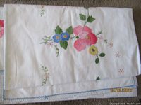 White tablecloth featuring hand embroidered floral motifs in pink, blue, yellow and green colors on white fabric.