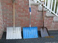Two snow shovels, one white metal blade and one blue plastic blade, both with handles, leaning against a brick wall accompanied by a brown coal shovel.