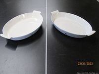 Two oval white interior with blue exterior enamel casserole dishes with tab handles shown from top view on dark surface.