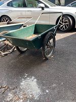 Side and front view of a green metal wheelbarrow cart with two spoked wheels on an asphalt surface, rust and dirt visible inside the cart.