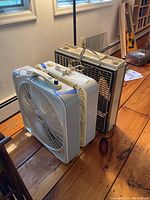 Photo showing three box fans with cords placed on wooden floor near wall.