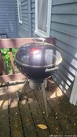 Black Weber charcoal kettle grill with lid, showing side and front views in outdoor setting. Grill stands on three legs with two wheels, front and vent controls visible.