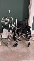 Two aluminum walkers and one manual wheelchair grouped against green wall on carpeted floor.