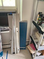 Mat cutter with adjustable rail and cutting mat leaning against wall near window and shelf.