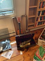 Acoustic guitar standing by window next to open saxophone case on wooden floor along with other boxes