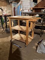 Wooden rolling kitchen cart seen from side angle showing top surface, shelves, wheels, and side handle with some utensils hanging.