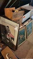 Three framed wall art pieces leaning against attic floor near cardboard boxes