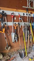 Wide view of garden and yard tools hanging on garage wall including shovels, rakes, trimmer, broom, root tool, pruners and grabber.
