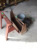 Wooden tool carrier with handle, red painted wooden sawhorse, galvanized metal bucket, cast iron pot, and some metal tools on the floor.