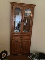 Full view of the wooden corner cabinet showing overall shape and structure, including glass doors, drawer, and lower cupboard doors.