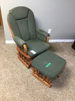 Photo showing front view of green fabric Dutailier glider rocker chair alongside matching ottoman on carpeted floor against beige wall.