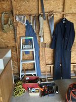 Photo of step ladder, wrenches, hand saws, coveralls hanging on the wall, and small toolbox on the floor with rope and crate with chainsaw chains.
