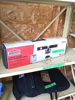 Boxed Job Mate laser level with adjustable tripod, showing front packaging and partially visible carrying case on shelf.