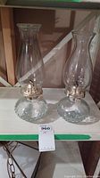Two clear glass oil lamps with round textured bases and mismatched chimneys on a shelf.