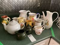 Group of various pitchers, creamers, teapots, and sugar bowls placed on a countertop showing the range of sizes and designs.