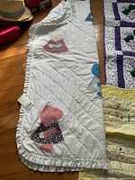 Vintage quilt with appliqué of girls wearing bonnets, showing white fabric with diagonal quilting lines and ruffled edging, detail of pink and floral fabric patches.