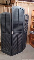 Front view of black three-panel wooden room divider made from window shutters, standing upright indoors in basement.