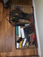 Overview photo showing various train cars, train engine, bridge parts, track and controller in a cardboard box on the wood floor.