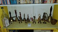 Photo showing a variety of brass and other types of bells arranged on a shelf with some wooden items and a bottle on the side.