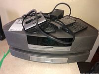 Front view of silver-gray Bose Wave Music System console on top of matching CD changer, display reads time, remotes and cables piled on top